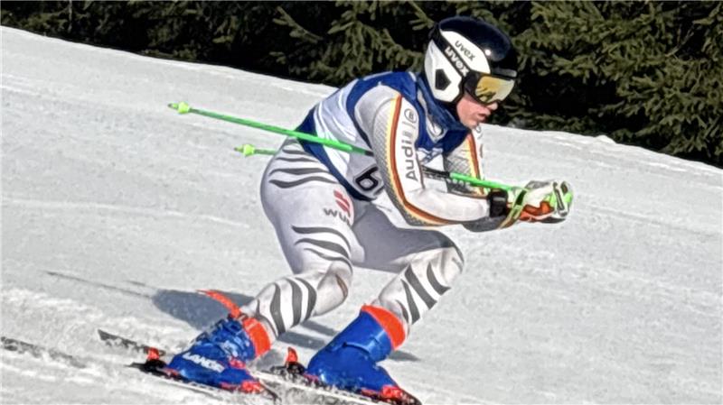 Skifahrer in Rennanzug mit Helm und Skistöcken in gebeugter Haltung auf schneebedeckter Piste vor Tannenwald