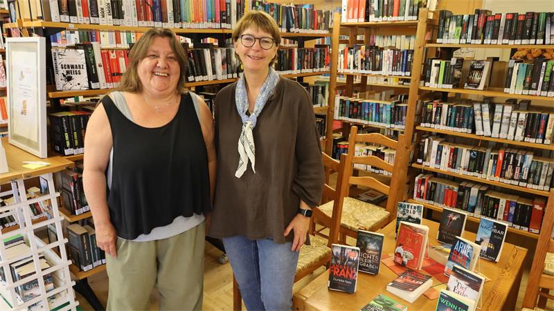 Zwei lächelnde Frauen stehen in einer Buchhandlung vor einem Tisch mit Büchern und Regalen voller Bücher im Hintergrund