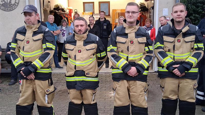 Vier Feuerwehrmänner in beigen Schutzanzügen mit reflektierenden Streifen stehen nebeneinander vor einer Gruppe von Menschen