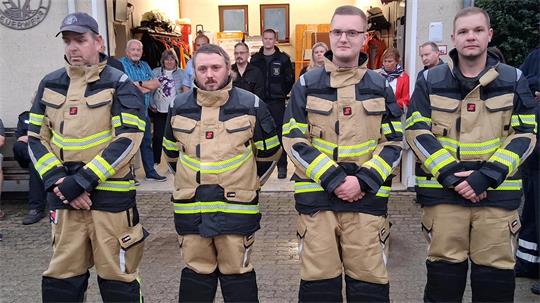 Vier Feuerwehrmänner in beigen Schutzanzügen mit reflektierenden Streifen stehen nebeneinander vor einer Gruppe von Menschen
