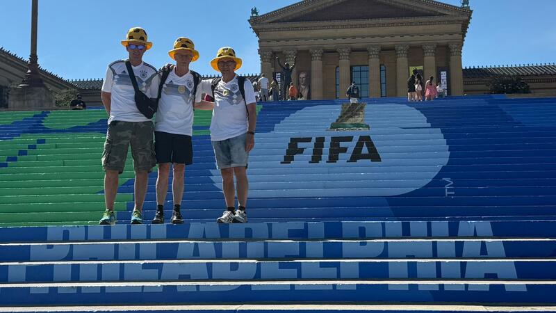 Drei Personen mit gelben Hüten stehen auf einer großen Treppe mit der Aufschrift 'PHILADELPHIA' und einem FIFA-Logo vor einem klassizistischen Gebäude.