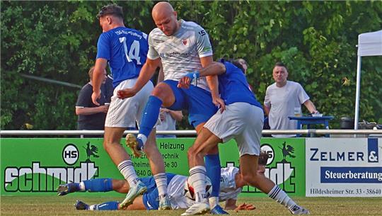 Drei Fußballspieler in blauen und weißen Trikots kämpfen um den Ball auf dem Spielfeld, im Hintergrund Werbetafeln und zwei Zuschauer an einem Tisch