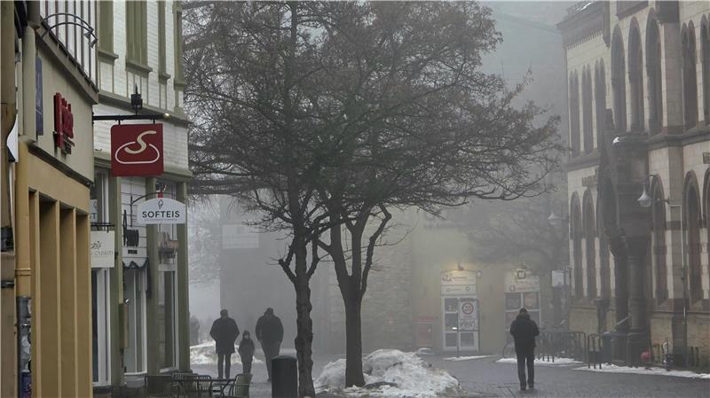 Zwei Personen gehen auf einer nebligen Kopfsteinpflasterstraße zwischen Fachwerkhäusern und kahlen Bäumen