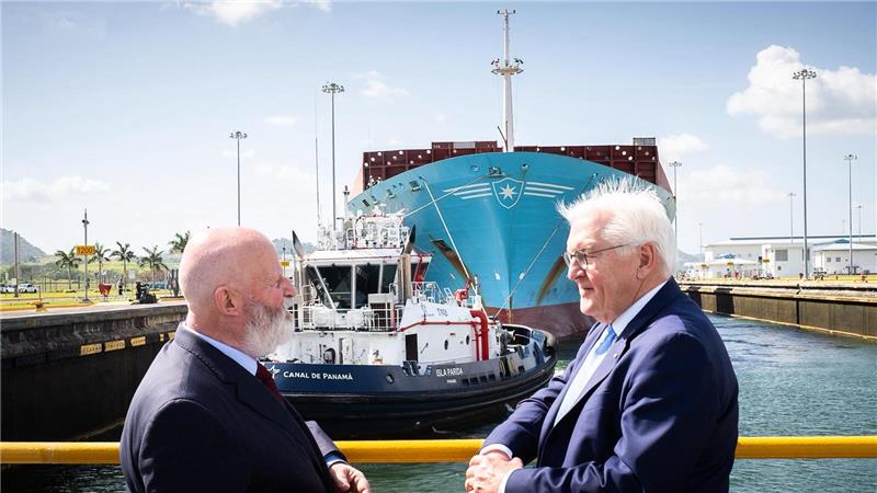 Zwei Männer in Anzügen stehen an einem Geländer vor einem großen Frachtschiff und einem Schlepper im Hafen