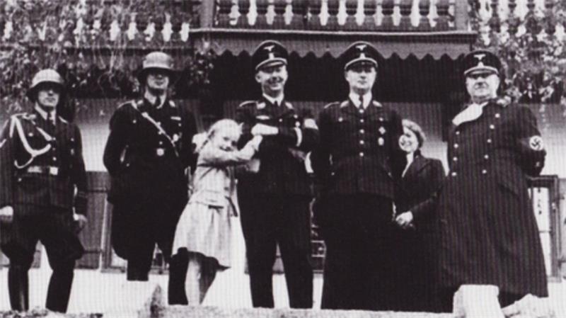 Fünf Männer in historischen Uniformen und zwei Kinder stehen vor einem Gebäude mit Balkon und Geländer