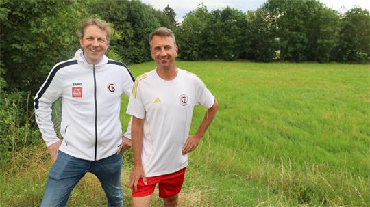 Zwei Männer in Sportkleidung stehen auf einer Wiese vor Büschen und Bäumen, einer trägt eine weiße Jacke und Jeans, der andere ein weißes T-Shirt und rote Shorts