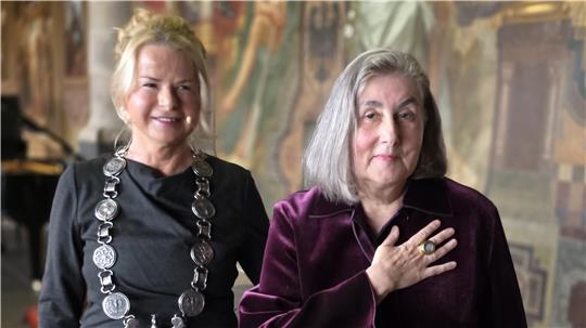 Das Foto zeigt die Kaiserringträgerin Katharina Fritsch auf der rechten Seite mit Oberbürgermeisterin Urte Schwerdtner auf der linken Seite. Katharina Fritsch präsentiert den Kaiserring an ihrer rechten Hand.