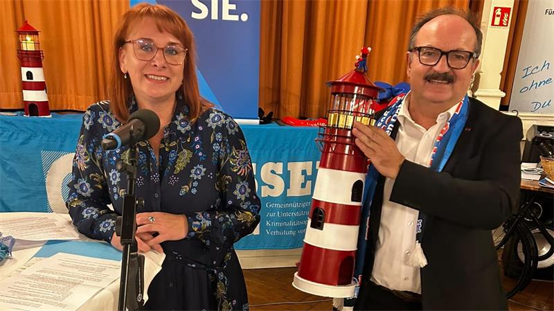 Günter Koschig übergibt nach 41 Jahren an Kristina Winter Zwei Personen stehen vor einem blauen Banner mit der Aufschrift 'WIR SEHEN SIE', die Frau trägt ein geblümtes Kleid und steht hinter einem Mikrofon, der Mann hält ein Modell eines rot-weiß gestreiften Leuchtturms