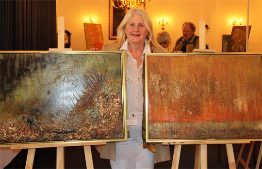 Eine blonde Frau steht hinter zwei Gemälden in einer Kunstausstellung. Im Hintergrund weitere Bilder und zwei Besucher.