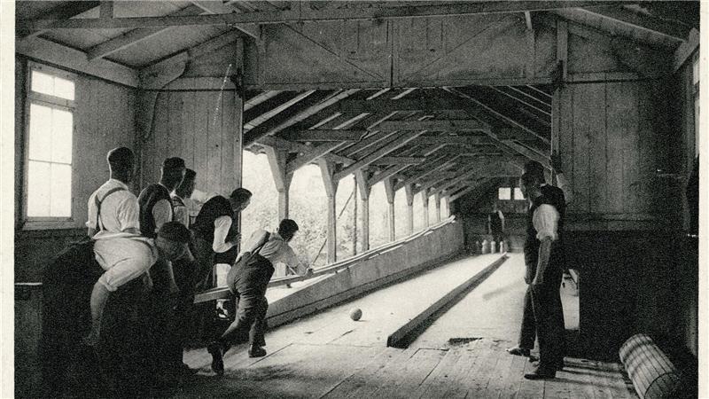 Gruppe von Männern in historischer Kleidung spielt Kegeln in einer langen Halle mit Holzbalken und großen Fenstern