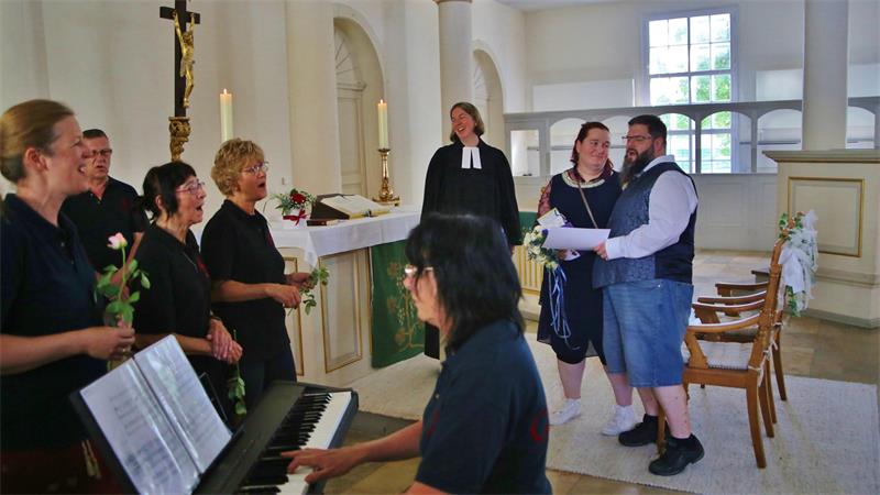 Das Foto zeigt einen Chor, eine Frau am Klavier, eine Pastorin und ein Ehrpaar in der Kirche.