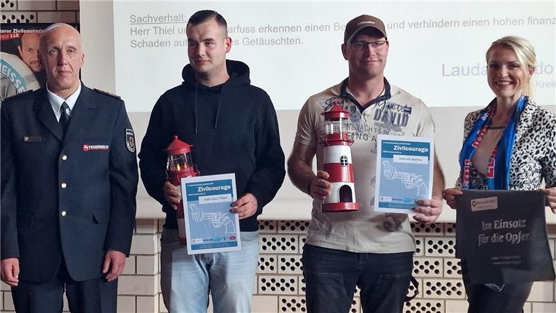 Vier Personen stehen nebeneinander in einem Raum, zwei halten Urkunden und Feuerlöscher, eine trägt eine Tasche mit Aufschrift