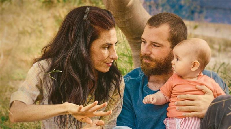 Drei Personen sitzen im Gras, eine Frau gestikuliert mit den Händen, ein Mann hält ein Baby auf dem Schoß, im Hintergrund Bäume und Natur, darunter der Filmtitel 'SORDA DER KLANG DER WELT' und die Namen 'MIRIAM GARLO' und 'ÁLVARO CERVANTES'.