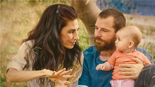 Drei Personen sitzen im Gras, eine Frau gestikuliert mit den Händen, ein Mann hält ein Baby auf dem Schoß, im Hintergrund Bäume und Natur, darunter der Filmtitel 'SORDA DER KLANG DER WELT' und die Namen 'MIRIAM GARLO' und 'ÁLVARO CERVANTES'.
