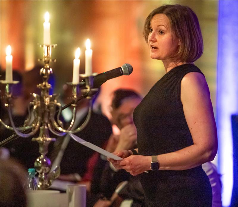 Frau in schwarzem ärmellosem Kleid hält ein Blatt Papier und spricht in ein Mikrofon vor einem goldfarbenen Kerzenleuchter mit brennenden Kerzen