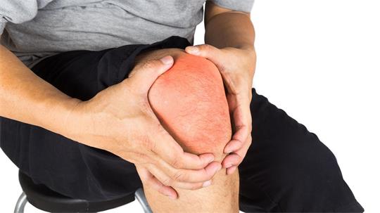 Ein Mann sitzt auf einem Stuhl und fasst sich mit beiden Händen an sein deutlich gerötetes gebeugtes Knie.