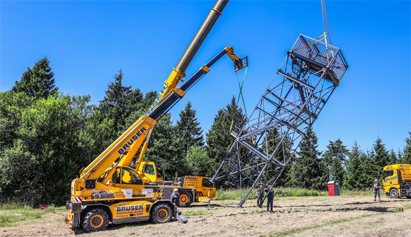 Zwei gelbe Autokräne heben einen großen Stahlgitterturm auf einer Wiese an, mehrere Personen stehen daneben.