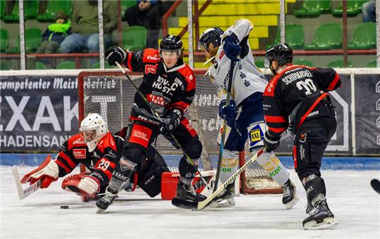 Eishockeyszene mit Torwart und mehreren Spielern in Aktion vor dem Tor auf dem Eis in einer Halle mit grünen Sitzreihen