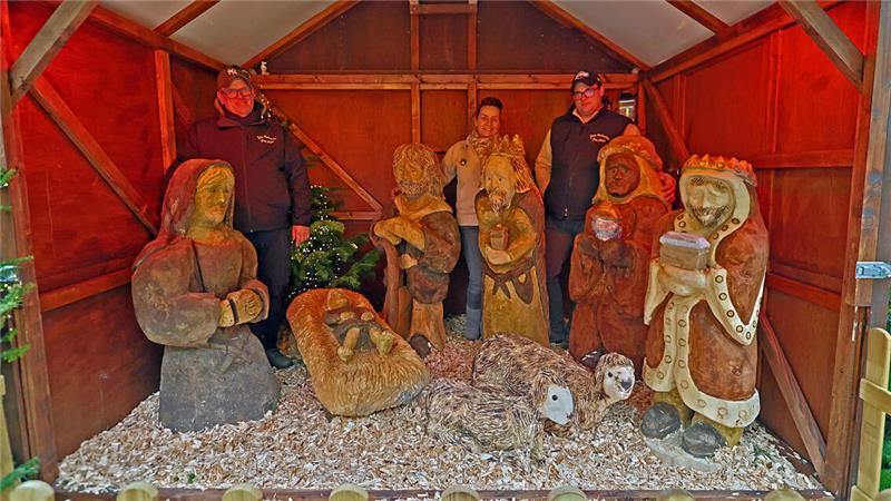 Holzfiguren in einer Weihnachtskrippe, darunter Maria, Josef, das Jesuskind in einer Krippe, drei Heilige Könige in einer Holzhütte. Dahinter stehen zwei Männer und eine Frau.