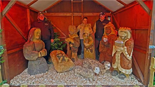 Holzfiguren in einer Weihnachtskrippe, darunter Maria, Josef, das Jesuskind in einer Krippe, drei Heilige Könige in einer Holzhütte. Dahinter stehen zwei Männer und eine Frau.