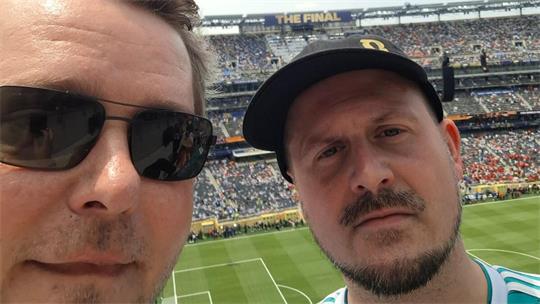 Zwei Männer mit Sonnenbrille und Baseballkappe vor einem vollen Fußballstadion bei Tageslicht