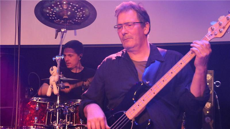 Ein Musiker spielt E-Bass auf einer Bühne, im Hintergrund ist ein Drummer an einem Schlagzeug mit einem kleinen Plüschtier darauf zu sehen.