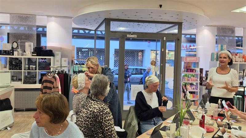 Mehrere Frauen unterschiedlichen Alters sitzen und stehen in einem hellen Raum mit Regalen und Tischen, auf denen Kosmetikprodukte stehen.