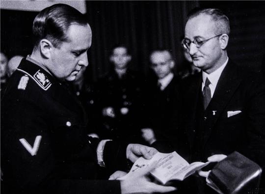 Zwei Männer in Anzügen und Uniformen betrachten gemeinsam ein gefaltetes Blatt Papier.