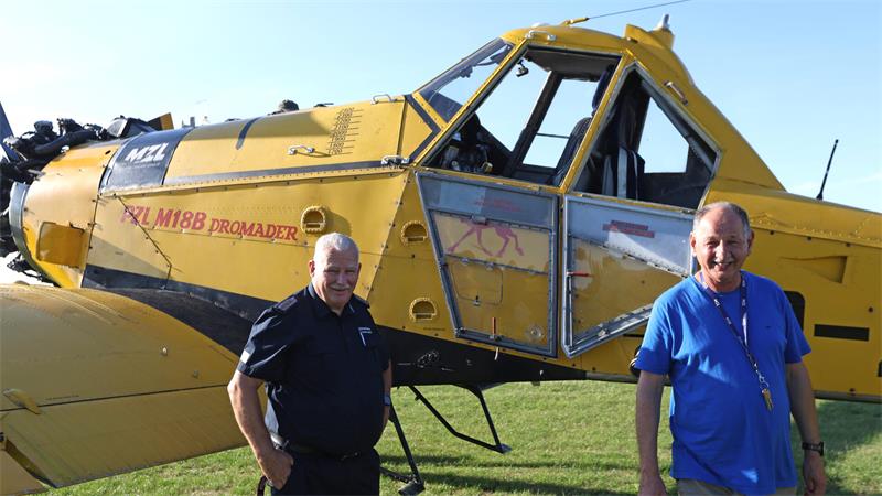 Ein gelbes Flugzeug ist zu sehen, einmotorig. Davor stehen zwei Männer.
