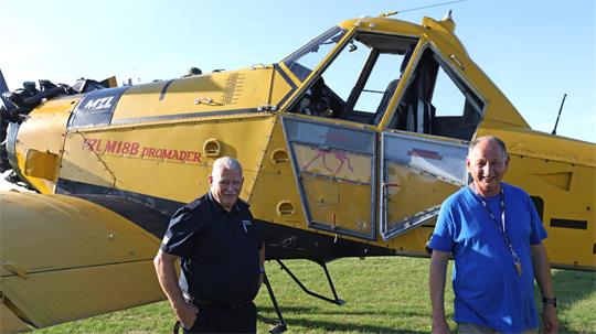 Ein gelbes Flugzeug ist zu sehen, einmotorig. Davor stehen zwei Männer.