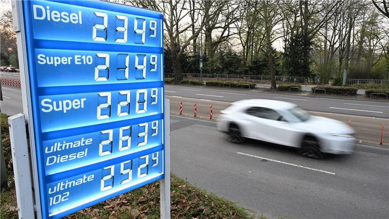 Auf dem Foto ist links eine blaue Anzeigentafel einer Tankstelle zu sehen mit den Spritpreisen. Rechts fährt auf der Straße ein Auto vorbei.