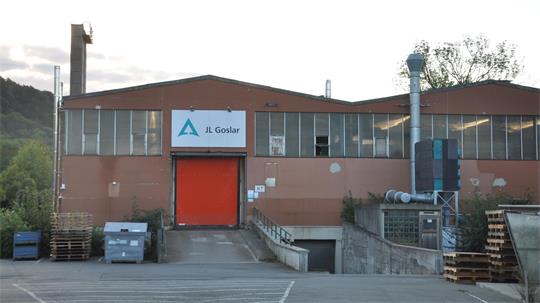 Industriegebäude mit roter Tür und Schild 'JL Goslar' über dem Eingang, umgeben von Paletten und Containern