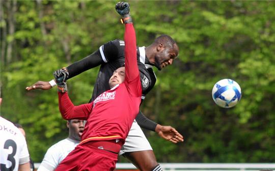 Fußballtorwart in rotem Trikot springt zum Ball, während ein Spieler in schwarzem Trikot und mehrere Spieler in weißen Trikots in Strafraumnähe stehen.