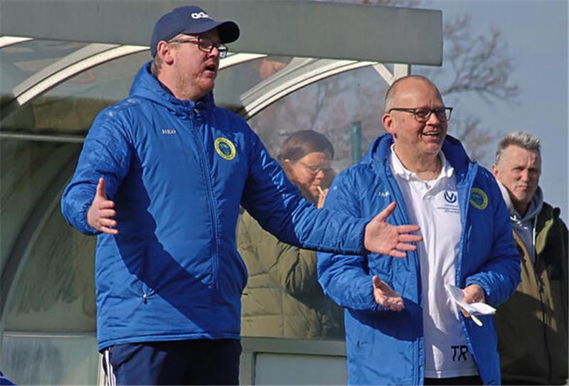 Trainer in blauer Jacke gestikuliert, ein zweiter Trainer in blauer Jacke lacht.