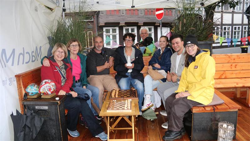 Gruppe von acht Personen sitzt auf Holzbänken um einen kleinen Tisch mit Schachbrett in einem überdachten Außenbereich.