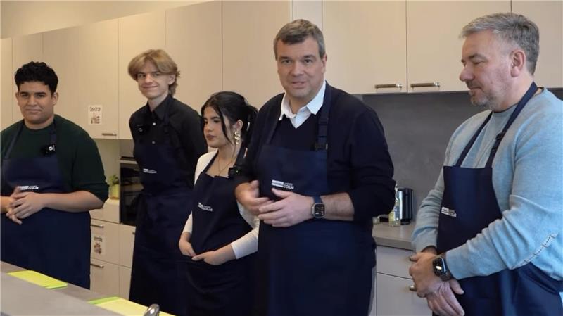 Fünf Personen mit Schürzen stehen in einer Küchenzeile. Vier hören zu, wie einer spricht.