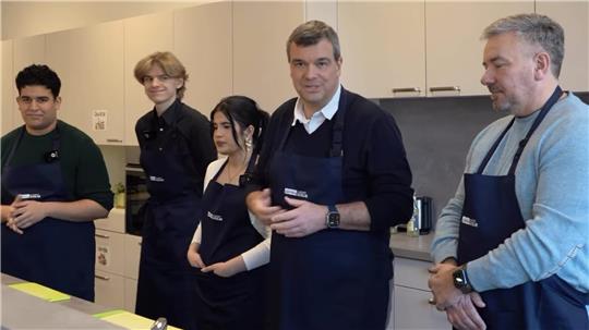 Fünf Personen mit Schürzen stehen in einer Küchenzeile. Vier hören zu, wie einer spricht.