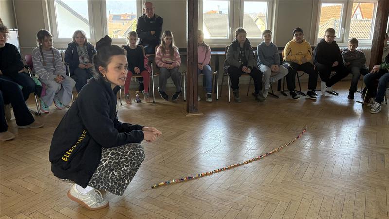 Gruppe von Kindern sitzt im Halbkreis auf Stühlen in einem Raum mit Holzboden, eine Person hockt in der Mitte vor einer auf dem Boden liegenden Schnur mit bunten Perlen.