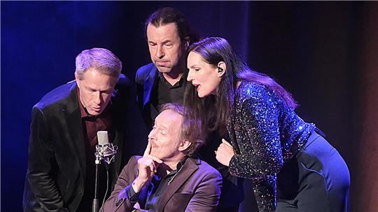 Drei Männer und eine Frau bilden ein Ensemble auf der Bühne. Sie singen in ein Mikro, ein Mann sitzt in der Mitte auf einem Stuhl.