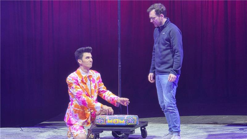 Osterfeld: Nach Neptuns Catwalk geht es zum Unterwasserball Zwei Männer auf einer Bühne vor rotem Vorhang, einer kniet in einem bunten Anzug neben einem kleinen Wagen mit einem bunten Kasten, der andere steht in Jeans und Pullover