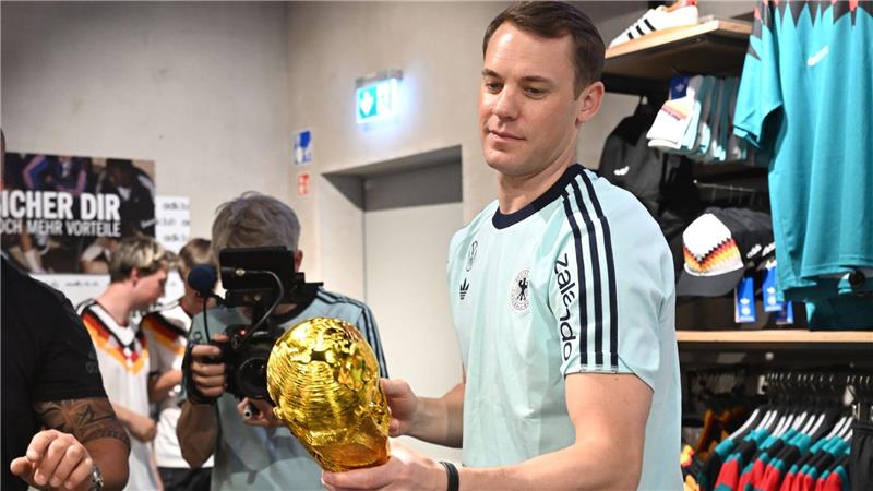 Lächelnd signiert Manuel Neuer einen goldenen WM-Pokal. 