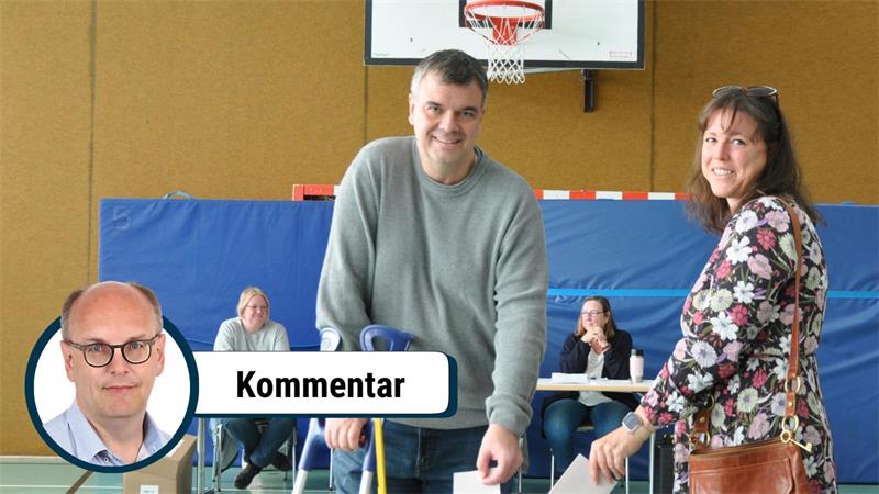 Zwei Personen werfen Stimmzettel in eine Wahlurne in einer Turnhalle, im Hintergrund sitzen zwei weitere Personen an einem Tisch