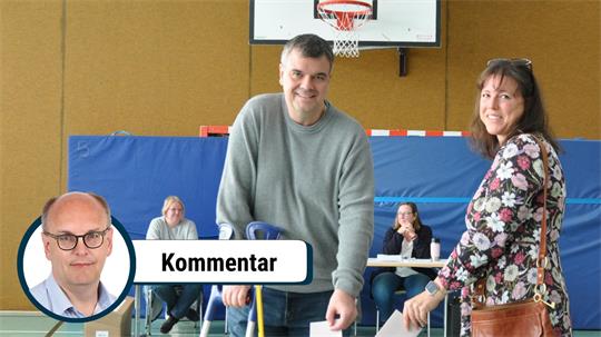 Zwei Personen werfen Stimmzettel in eine Wahlurne in einer Turnhalle, im Hintergrund sitzen zwei weitere Personen an einem Tisch