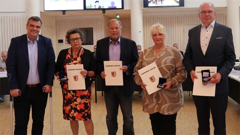 Fünf Personen stehen nebeneinander in einem hellen Raum, vier von ihnen halten Urkunden mit einem Wappen, drei Personen halten eine Medaille in einer Schachtel
