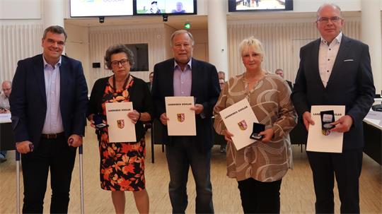 Fünf Personen stehen nebeneinander in einem hellen Raum, vier von ihnen halten Urkunden mit einem Wappen, drei Personen halten eine Medaille in einer Schachtel