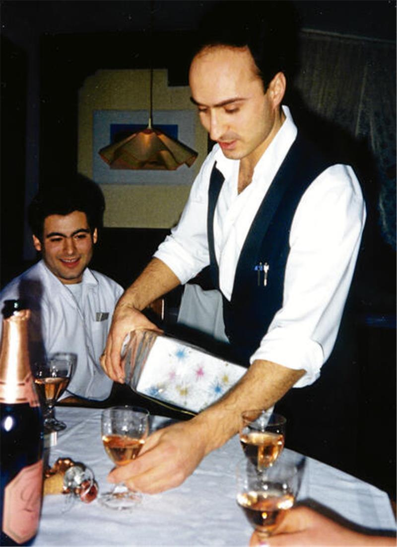 Mann in weißem Hemd und schwarzer Weste schenkt an gedecktem Tisch mit Sektflasche und Gläsern ein.