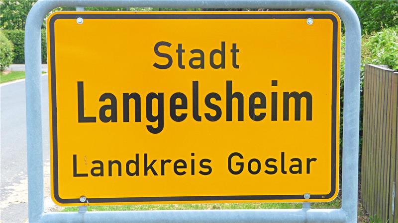 Gelbes Ortsschild mit schwarzer Schrift 'Stadt Langelsheim Landkreis Goslar' an einer Straße mit Grünflächen und Bäumen im Hintergrund.