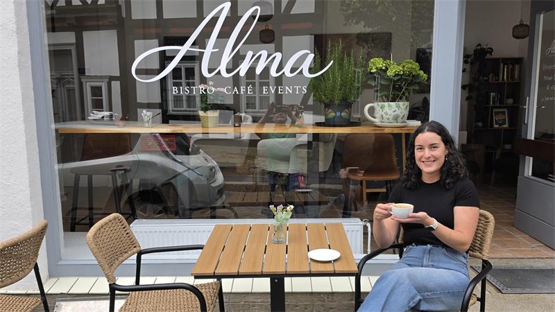 Frau sitzt mit Tasse vor einem Bistro-Café namens Alma an einem Holztisch mit Stühlen aus Korbgeflecht und Metallrahmen.