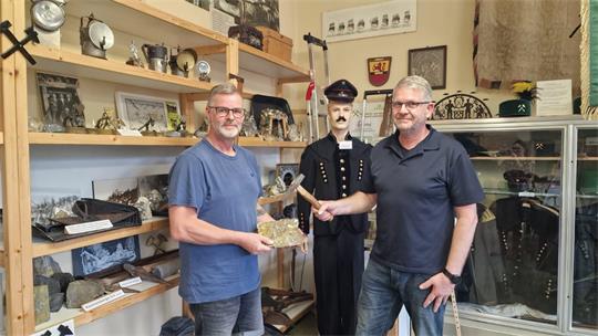 Zwei Männer stehen in einem Raum mit Regalen voller alter Gegenstände und schütteln sich die Hände, zwischen ihnen eine Schaufensterpuppe in historischer Uniform.