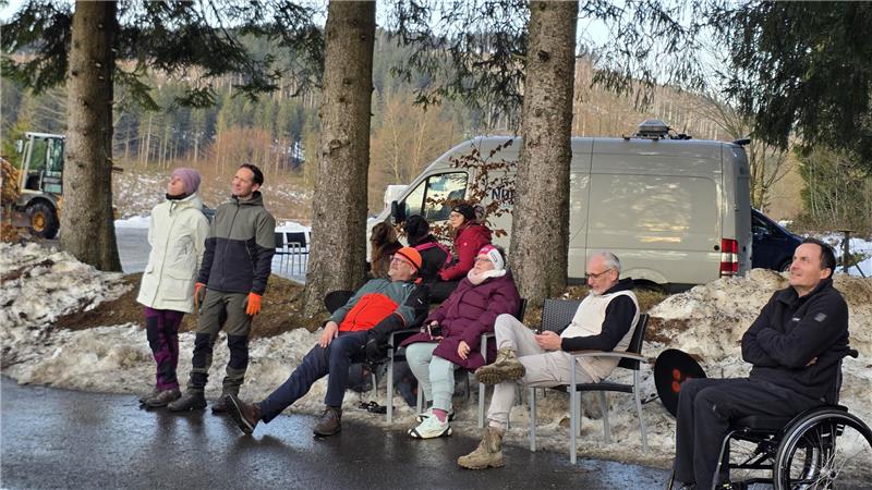 Gruppe von Menschen in Winterkleidung steht und sitzt auf einer Bank am Straßenrand vor einem weißen Transporter in einer verschneiten Waldlandschaft.